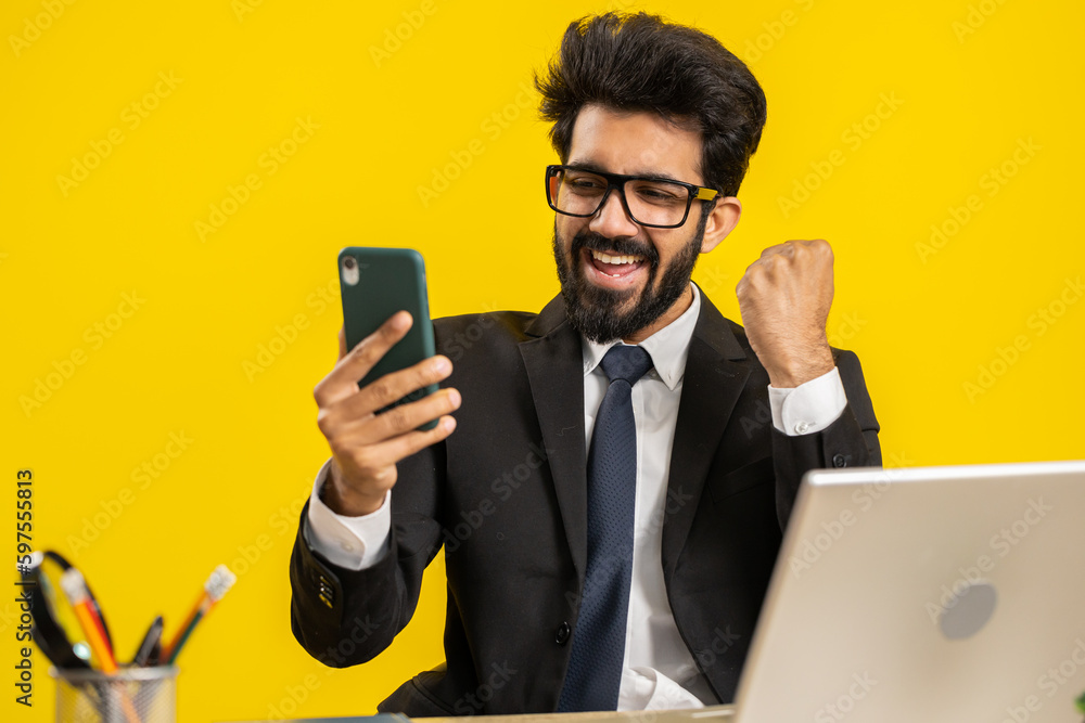 Happy amazed indian businessman working on smartphone shocked by sudden victory game winning lottery jackpot achievemen good career news. Excited hindu man freelancer at office on yellow background Happy amazed indian businessman working on smartphone shocked by sudden victory game winning lottery jackpot achievemen good career news. Excited hindu man freelancer at office on yellow background