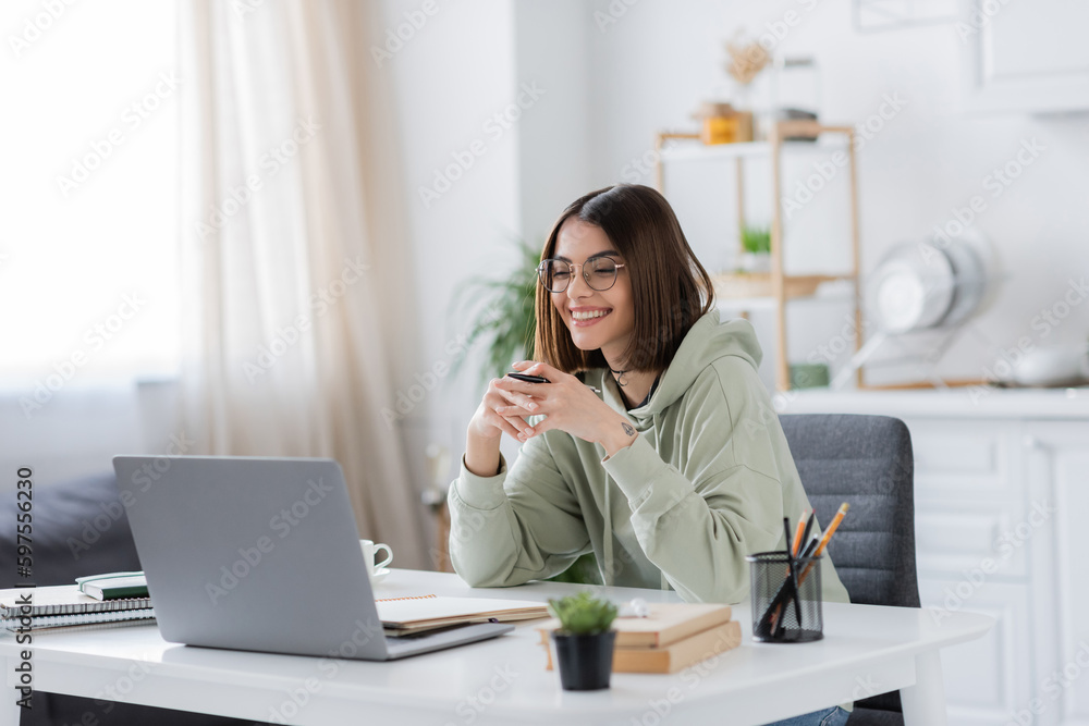 Smiling freelancer in eyeglasses holding pen near laptop and notebooks at home. Smiling freelancer in eyeglasses holding pen near laptop and notebooks at home.