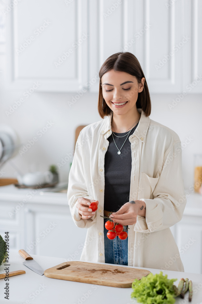 Positive woman holding cherry tomatoes near vegetables and cutting board in kitchen. Positive woman holding cherry tomatoes near vegetables and cutting board in kitchen.