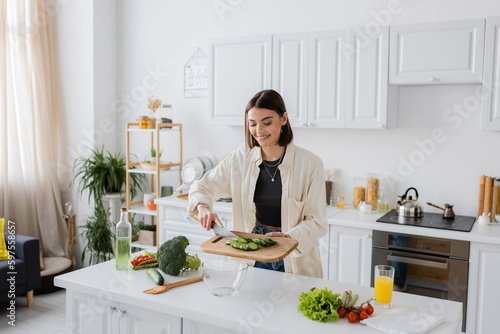 Cheerful woman holding cut cucumber on cutting board while cooking salad in kitchen.