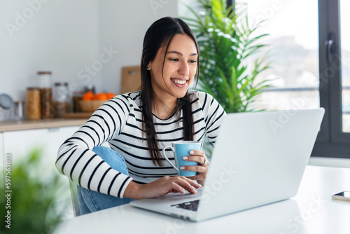 Foto Smiling young woman doing video call with laptop while holding a cup of coffee in the kitchen at home