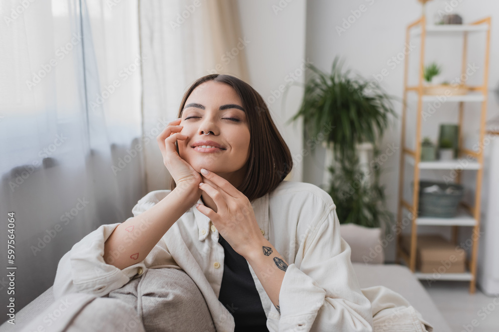 Overjoyed tattooed woman touching face while sitting on couch at home. Overjoyed tattooed woman touching face while sitting on couch at home.