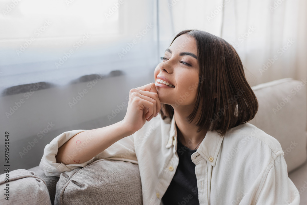 Cheerful young woman in shirt looking away while sitting on couch at home. Cheerful young woman in shirt looking away while sitting on couch at home.