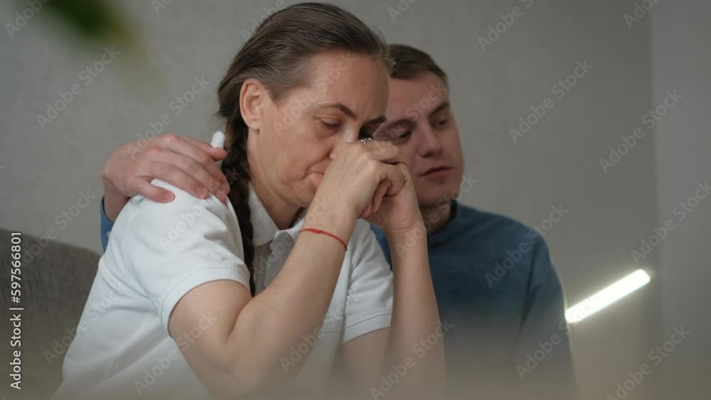 The son calms and tries to cheer up his mother while sitting on the sofa nearby The son calms and tries to cheer up his mother while sitting on the sofa nearby