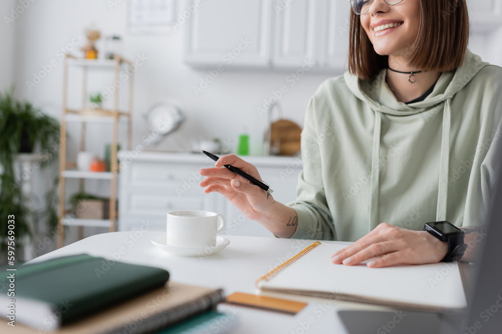Cropped view of positive freelancer holding pen near coffee and notebooks at home. Cropped view of positive freelancer holding pen near coffee and notebooks at home.
