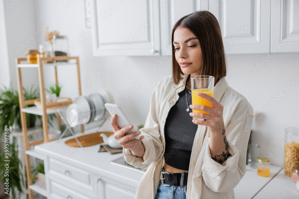 Brunette woman holding orange juice and using smartphone in kitchen.