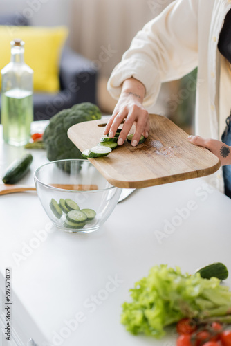 Cropped view of tattooed woman adding sliced cucumber in bowl in kitchen.