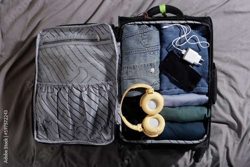 carry-on suitcase with blue clothes and large headphones, charger and cell phone on the bed