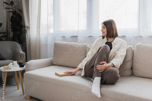 Smiling young woman taking book while sitting on couch at home.