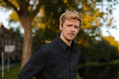 Young fun man wear black shirt and t-shirt walking rest relax in spring or summer green city park go down alley sunshine lawn outdoors on nature. Urban lifestyle leisure concept. Blonde man look calm.