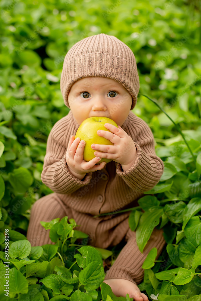 a little boy with big eyes in a brown jumpsuit and a hat is sitting in the green grass and holding a green apple to his mouth a little boy with big eyes in a brown jumpsuit and a hat is sitting in the green grass and holding a green apple to his mouth