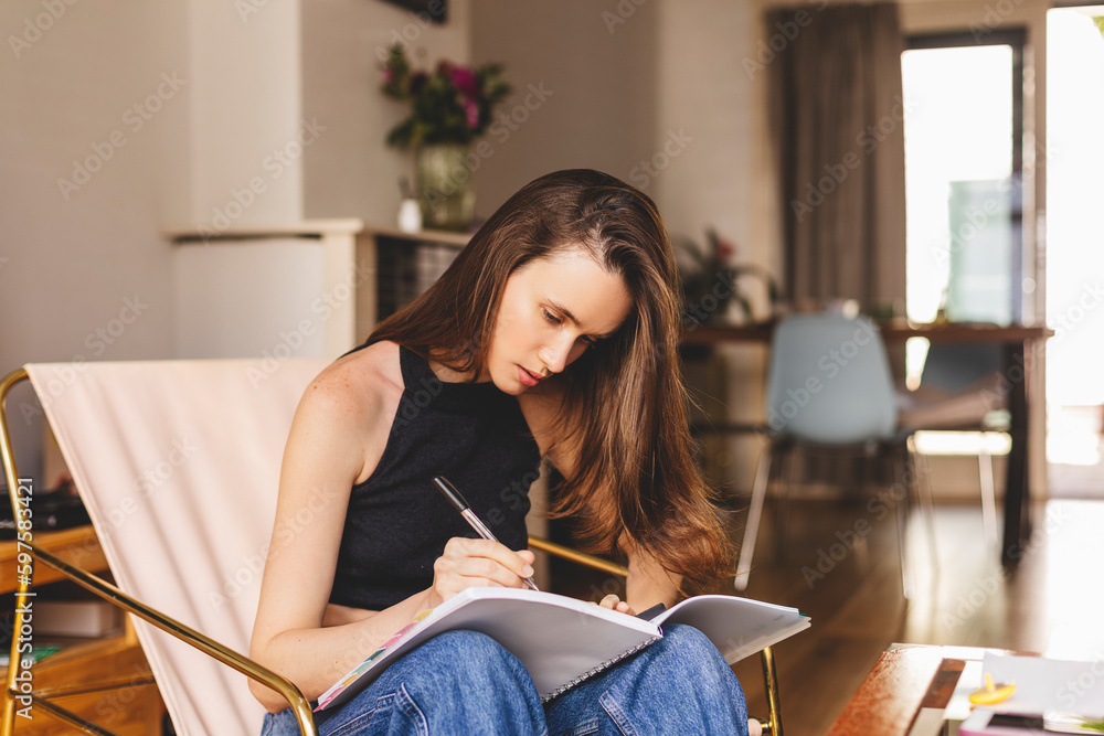 Beautiful young woman writing something in the note pad while sitting on the sofa at living room at home, charming female studying doing homework at home, girl writes diary or work at house. Beautiful young woman writing something in the note pad while sitting on the sofa at living room at home, charming female studying doing homework at home, girl writes diary or work at house.