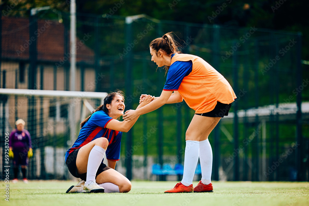 Happy female player helps her teammate to get up on soccer pitch.