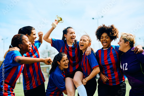 Cheerful soccer plyer holds winning trophy while being carried by her teammates at stadium.