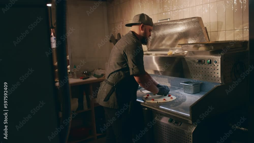 Medium side shot of Caucasian male chef making pizza alone in tiled dark restaurant kitchen, placing cherry tomatoes on dough base and pinching it for star shape Medium side shot of Caucasian male chef making pizza alone in tiled dark restaurant kitchen, placing cherry tomatoes on dough base and pinching it for star shape