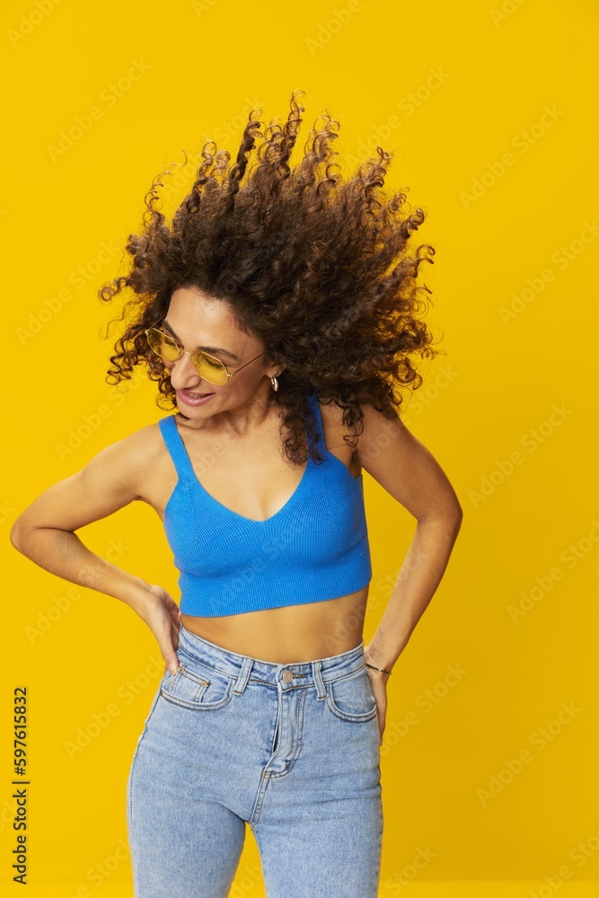 Woman with curly afro hair in a blue T-shirt on a yellow background dancing flying hair with sunglasses yellow, hand signs, look into the camera, smile with teeth and happiness, copy space Woman with curly afro hair in a blue T-shirt on a yellow background dancing flying hair with sunglasses yellow, hand signs, look into the camera, smile with teeth and happiness, copy space