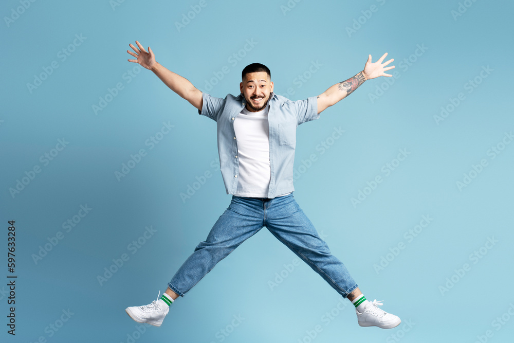 Funny smiling Asian man with stylish tattooed on arms jumping, having fun  isolated on blue background