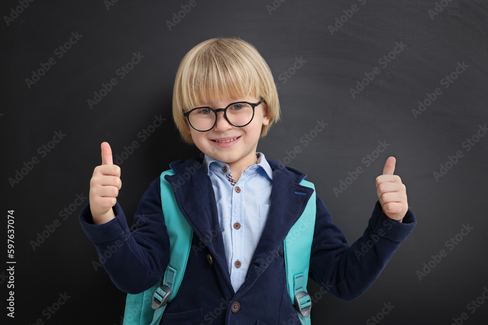 Happy little school child with backpack showing thumbs up near chalkboard Happy little school child with backpack showing thumbs up near chalkboard