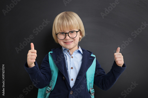 Happy little school child with backpack showing thumbs up near chalkboard