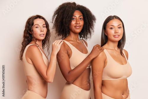 Three different women in beige underwear pose against white background put hands on shoulder of friend next to, comfort underclothes concept, copy space © South House Studio