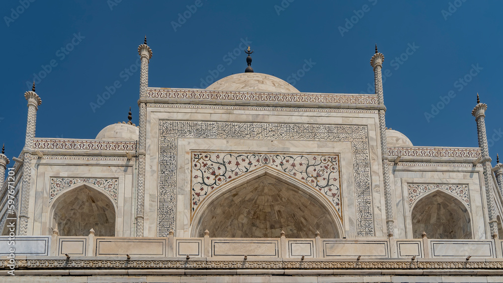 Details of the architecture of the famous Taj Mahal. The upper part of the white marble mausoleum with domes, spires, arches. There are ornaments on the walls, inlaid with precious stones. Blue sky.