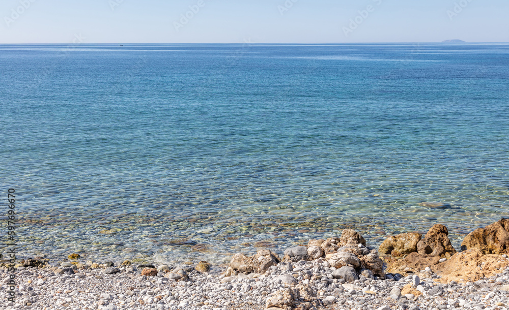 Rocky empty beach transparent calm sea water clear blue sky background. Greek seascape, sunny day.