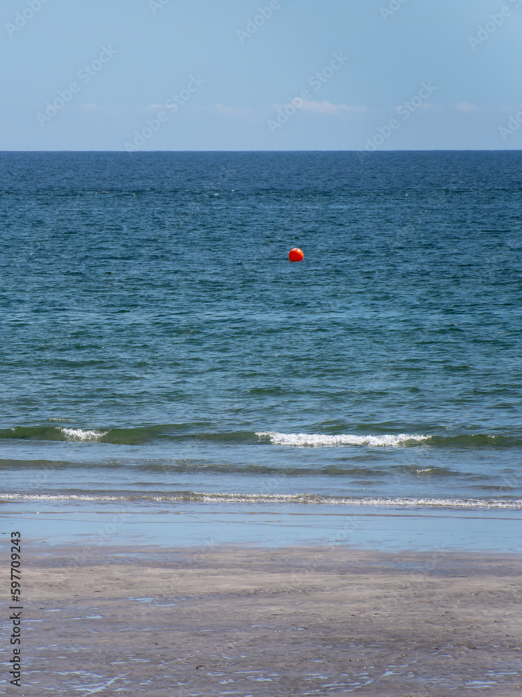 Blue sea water and a sandy beach under a blue sky. Orange buoy on the water surface. Red balloon floating on sea