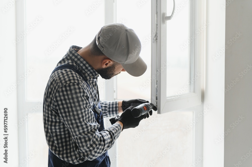 Workman in overalls installing or adjusting plastic windows in the living room at home Workman in overalls installing or adjusting plastic windows in the living room at home