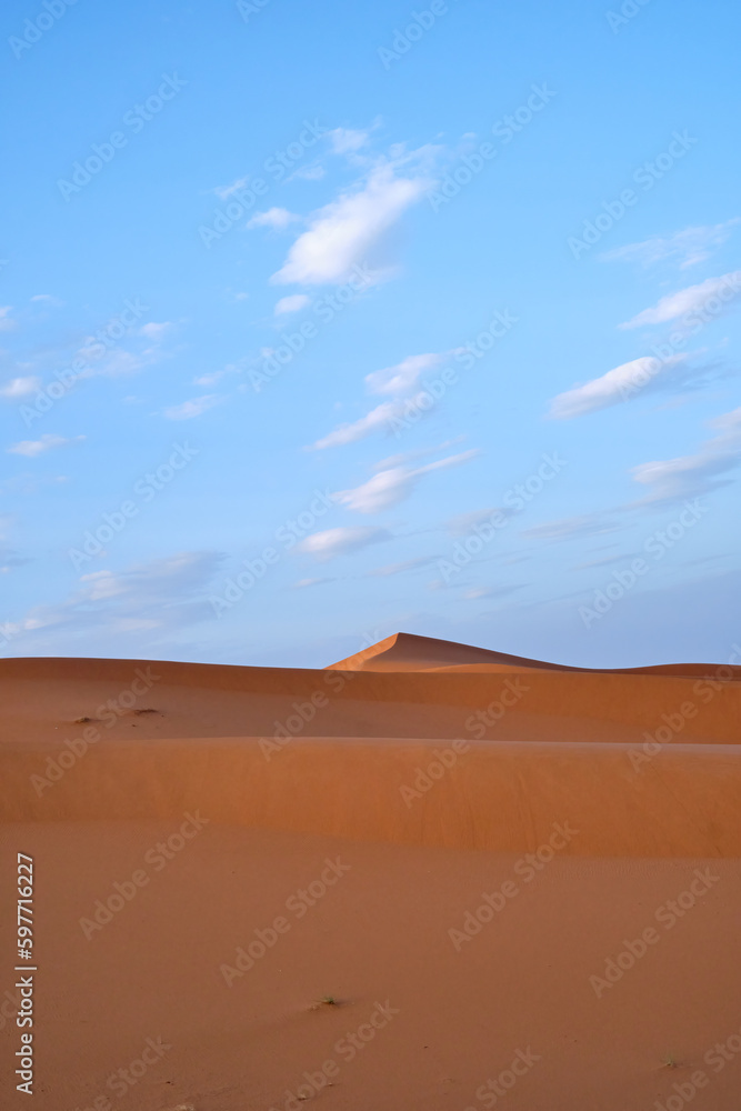 A portrait shot of the sand dunes in the Sahara desert, Morocco, on a blue sky day with clouds.