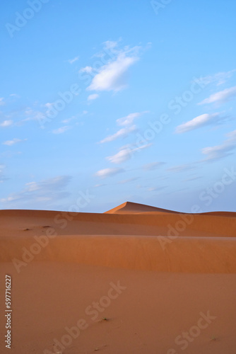 A portrait shot of the sand dunes in the Sahara desert, Morocco, on a blue sky day with clouds.