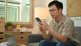 Image of adult asian man in casual watching video or playing game on smart phone, sitting on couch at home