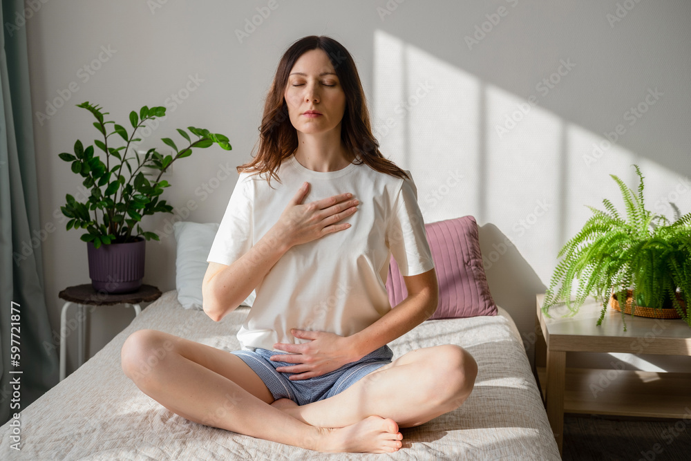 Pretty female sitting on bed in lotus posture with hands on her chest and belly doing pranayama techniques. Doing yoga at home. Self care and positive emotions. - obrazy, fototapety, plakaty 