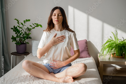 Pretty female sitting on bed in lotus posture with hands on her chest and belly doing pranayama techniques. Doing yoga at home. Self care and positive emotions. photo