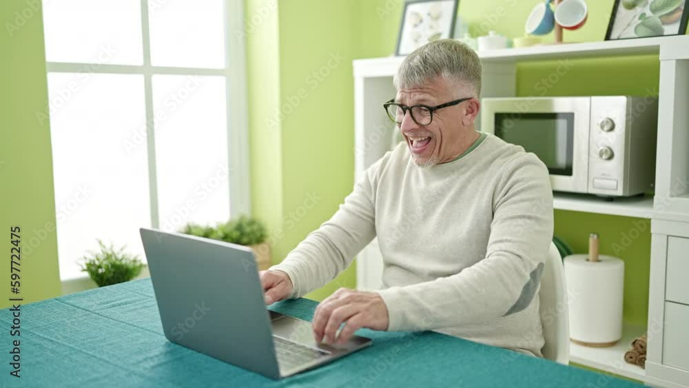 Middle age grey-haired man using laptop sitting on table dancing at home Middle age grey-haired man using laptop sitting on table dancing at home