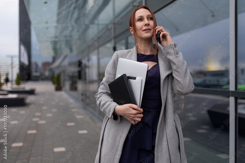 young caucasian woman manager speaks on the phone holding documents in her hand on the background of the building