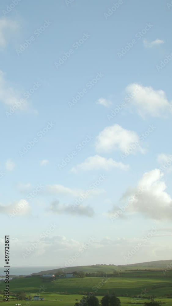 An image of fast-moving white cumulus clouds against a blue sky over green Irish hills was captured in a timelapse video. Vertical video.