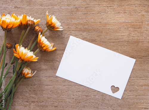 White card with a heart hole punch and orange chrysanthemum blooming on rustic wooden background. Template for Anniversary, valentine, wedding, and mother day. photo