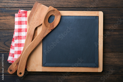 Cutting board with kitchen spoons and red napkin and parsley, tomato for cooking on old wooden dark background. Vegetarian food, health or cooking concept. Food background with free space for text.