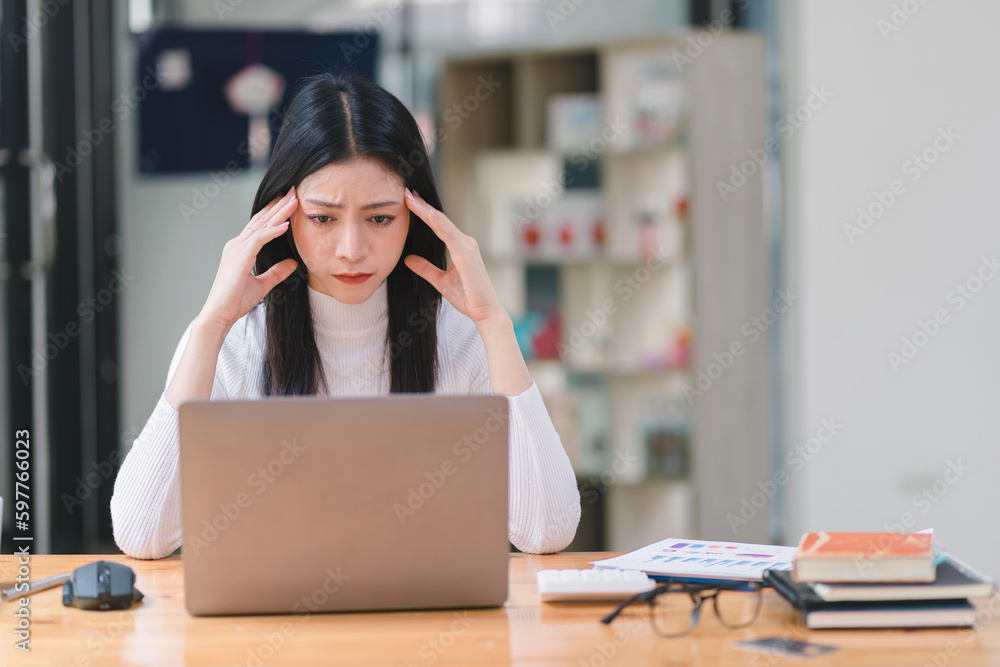The stressed and exhausted millennial Asian businesswoman is seen sitting at her office desk with her hand on her head, indicating a hard working day where she is overloaded with work. - obrazy, fototapety, plakaty 