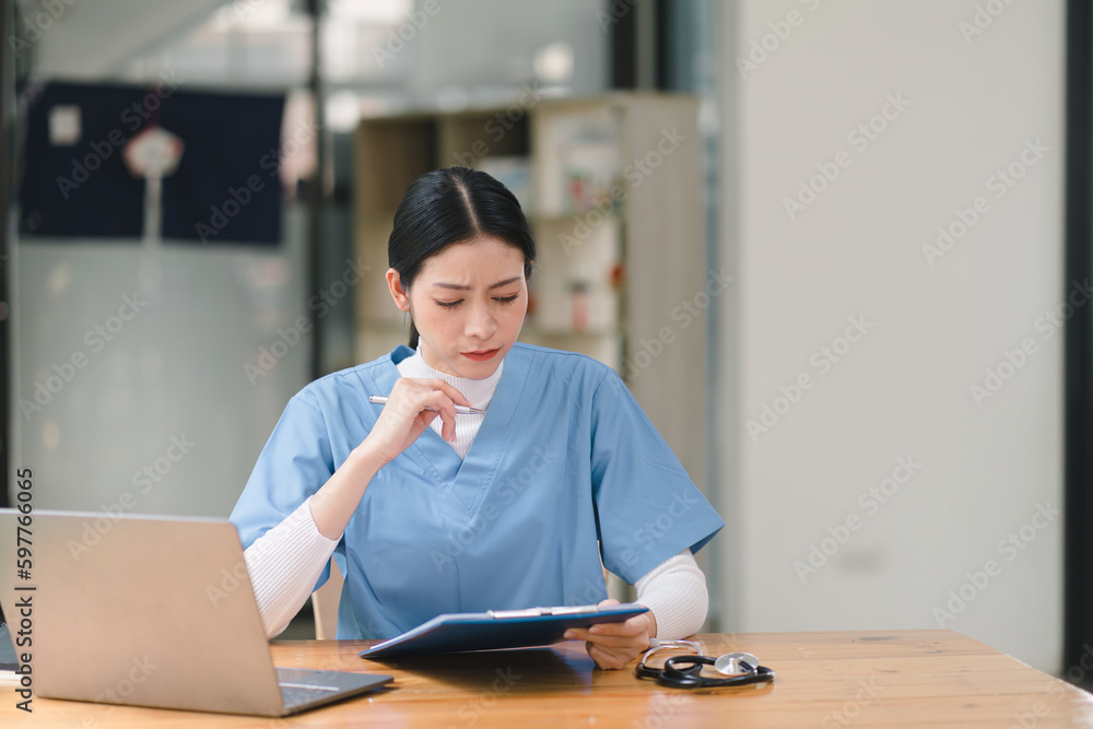 A concentrated female doctor or nurse is working online with a laptop while sitting at a desk in a hospital consultation room. medicine, technology and healthcare concept A concentrated female doctor or nurse is working online with a laptop while sitting at a desk in a hospital consultation room. medicine, technology and healthcare concept