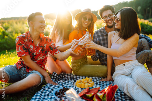 Cheerful company on sunny meadow at picnic is resting, drinking beer, cheers. Stylish friends enjoy sunny day on beautiful green meadow. Vacation, picnic, friendship or holliday concept.