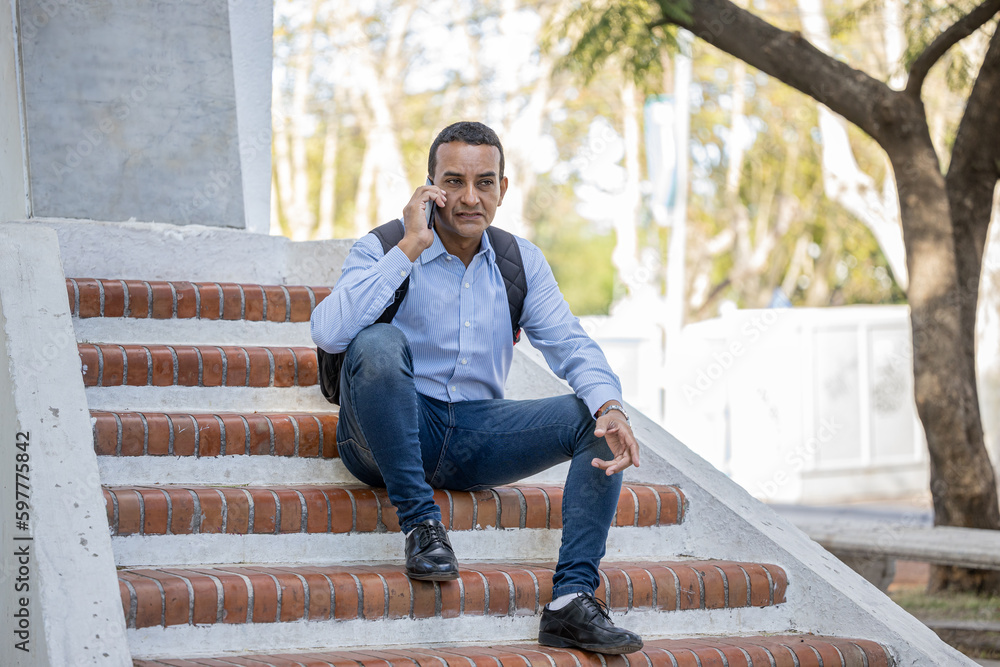 Fototapeta premium Young latino man sitting on some stairs stairs of a monument on his mobile phone.