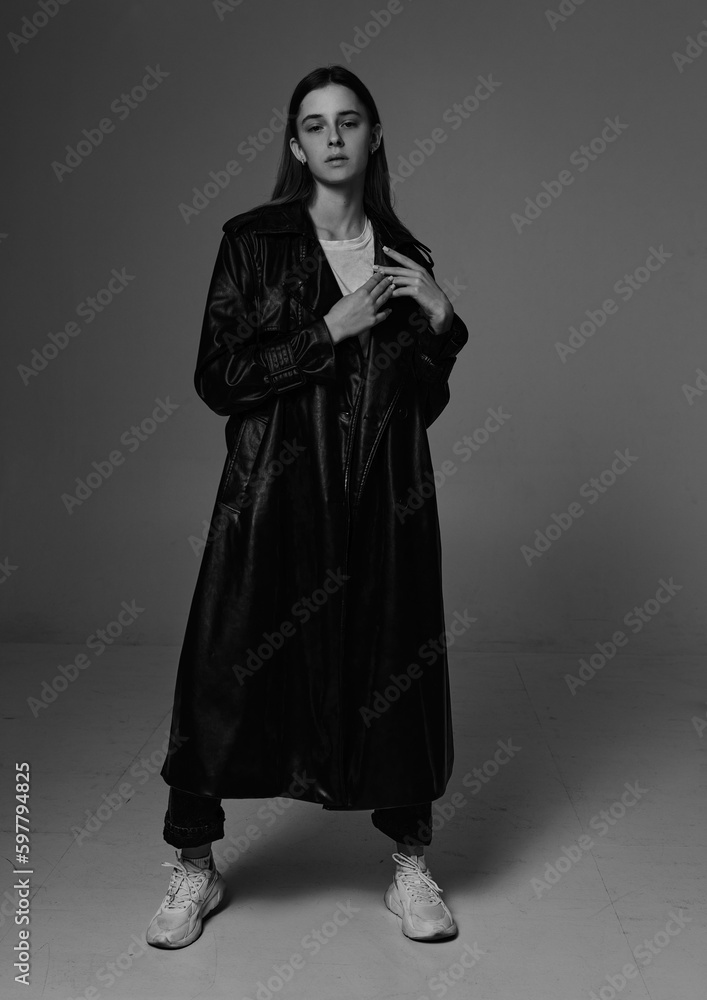 Young beautiful fashion model in long leather coat and dark jeans on dark grey background.