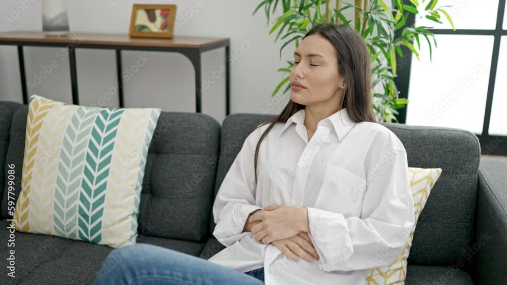 Young beautiful hispanic woman suffering for stomach ache sitting on sofa at home Young beautiful hispanic woman suffering for stomach ache sitting on sofa at home