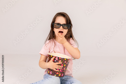 kid in black 3D glasses eating tasty popcorn in carton bucket on white background in light studio