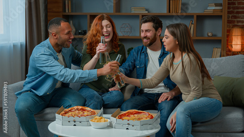 Happy multiracial friends celebrate festive event clink beer bottles eat pizza order food delivery sit at home sofa excited women and men laughing eating rest weekend friday party with snacks drinks