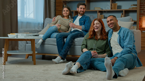 Multiracial friends men women watching television at home hispanic couple relax on couch african american caucasian family sit on floor enjoy TV comedy film. Diverse ethnic people laughing drink beer