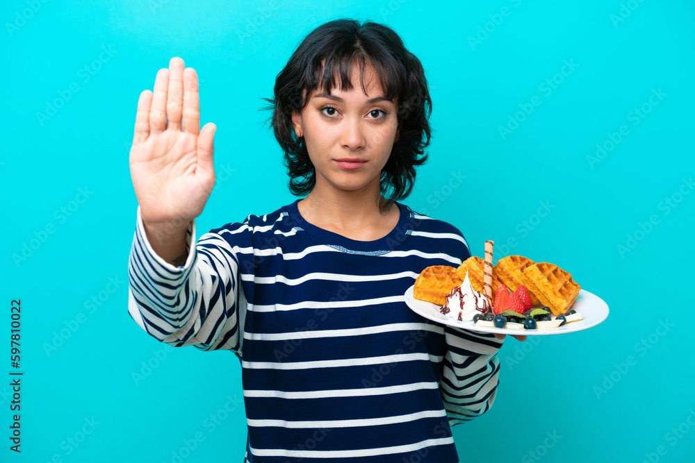 Young Argentinian woman holding waffles isolated on blue background making stop gesture Young Argentinian woman holding waffles isolated on blue background making stop gesture