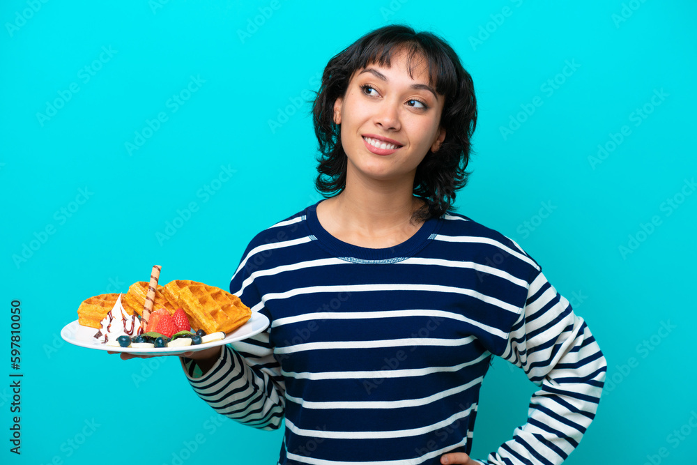 Young Argentinian woman holding waffles isolated on blue background thinking an idea while looking up