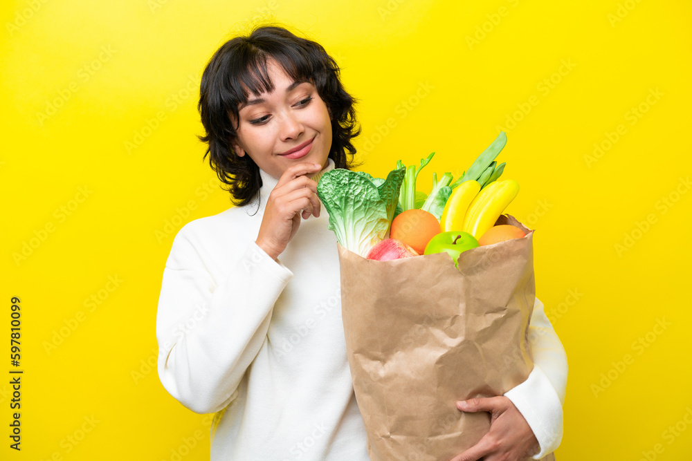 Young Argentinian woman holding a grocery shopping bag isolated on yellow background looking to the side and smiling Young Argentinian woman holding a grocery shopping bag isolated on yellow background looking to the side and smiling
